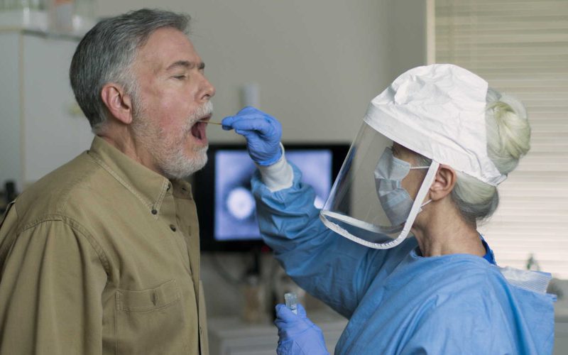 A mature Caucasian man in a clinical setting being swabbed by a healthcare worker in protective garb to determine if he has contracted the coronavirus or COVID-19.